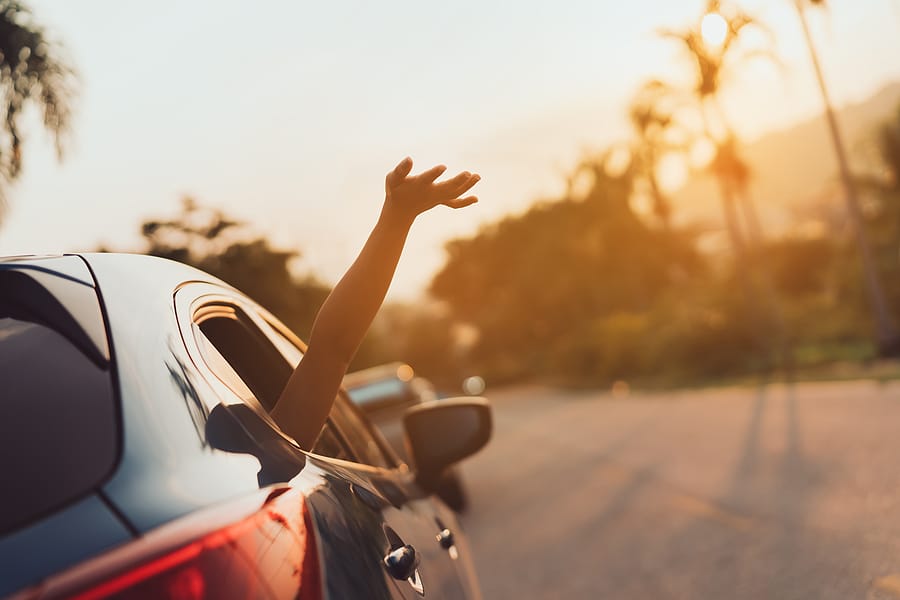 Girls happily travel with friends on a road trip. Rear view of hatchback vehicle with woman waving her right hand out the front-passenger window.